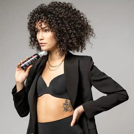 Egy nő göndör hajjal, fekete blézerben és melltartóban, aki egy göndör hajú terméket tart.