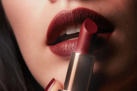 Egy nő nyitott szája sötét piros matt rúzzsal, miközben egy rózsaarany alappal rendelkező rúzstokát tart.