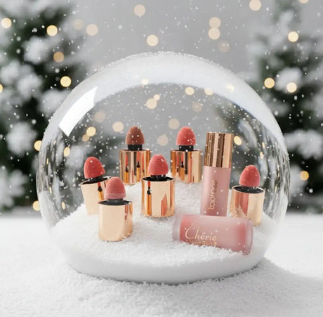Egy ünnepi hóglobusz miniatűr rózsa-arany rúzs tubusokkal és egy hozzá illő pirosítóval, havas ünnepi háttér előtt.